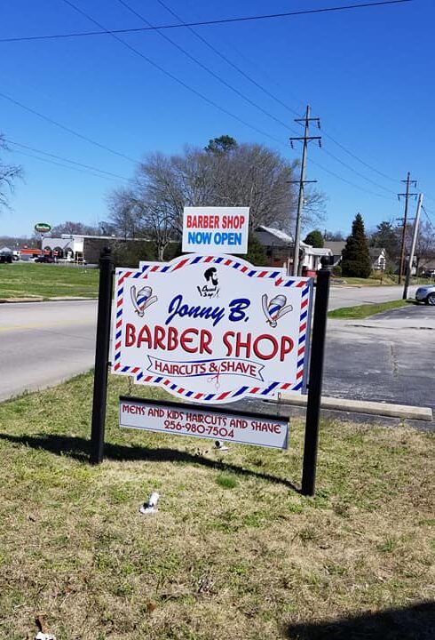 Sign for "Jonny B. Barber Shop" with a red, white, and blue striped border, advertising haircuts and shaves. A smaller sign reads "Barber Shop Now Open."