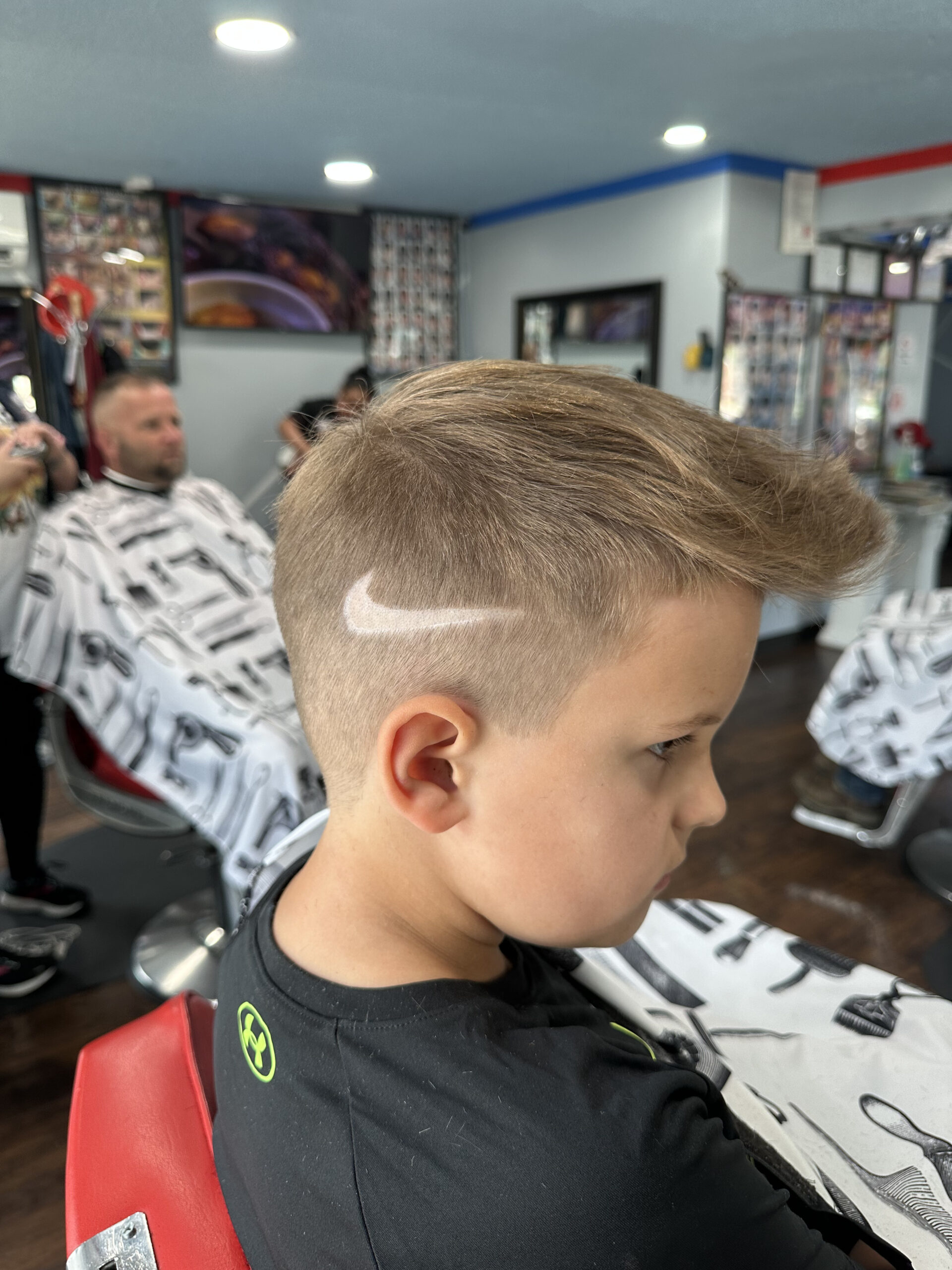 Young boy with light brown hair in a barber chair, showcasing a fade haircut with a white swoosh design on the side. The barber shop has a modern, colorful interior.