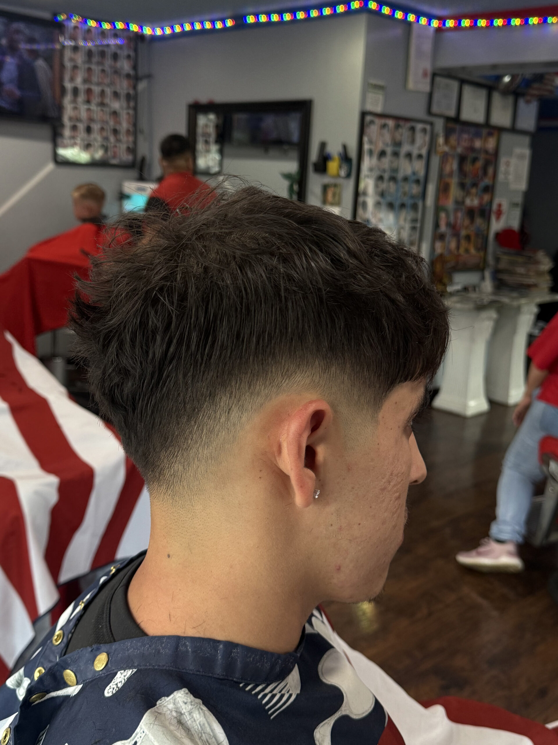 Young man with a fresh undercut hairstyle sits in a barbershop. The decor includes colorful lights and barber-themed posters, creating a lively atmosphere.