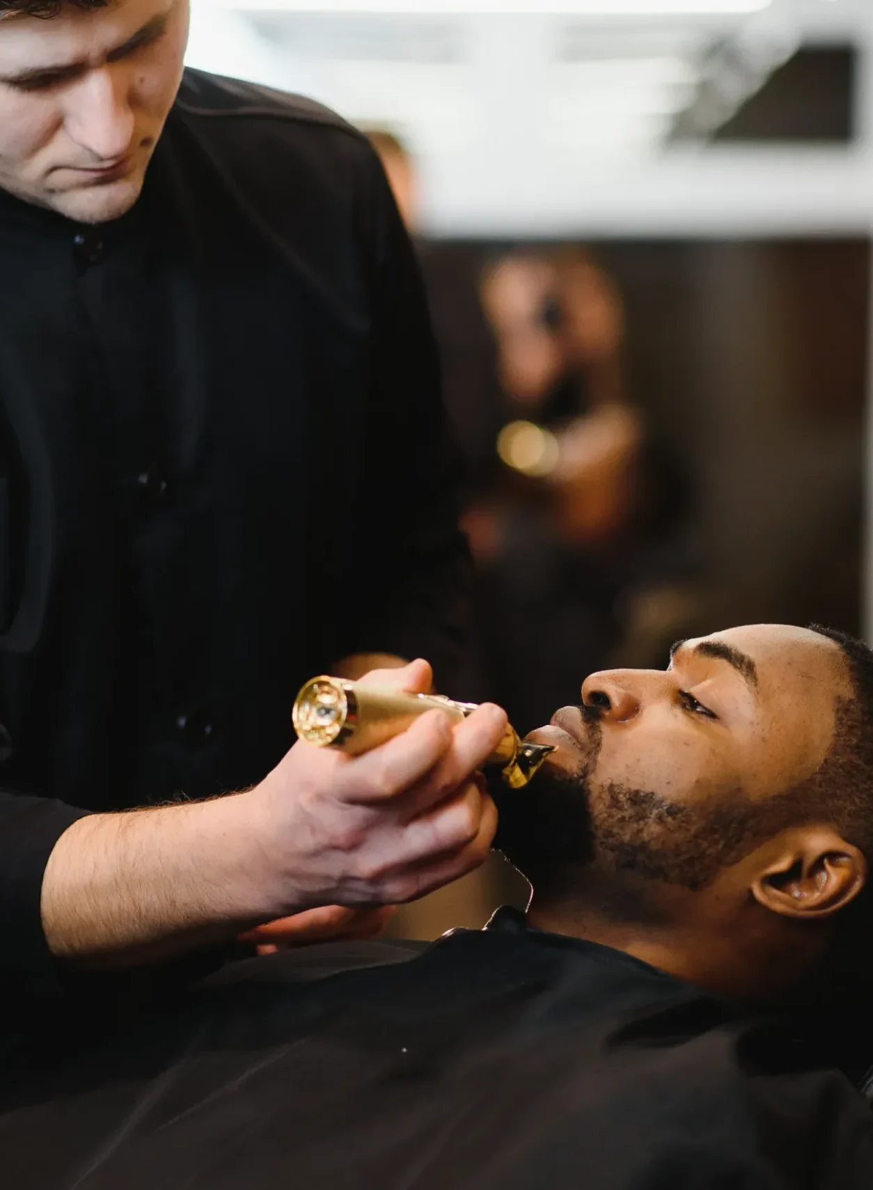 A barber wearing a black shirt trims a client's beard with an electric trimmer. The client is reclined and relaxed, conveying a calm atmosphere.